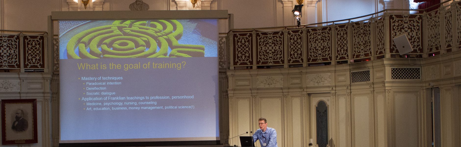 Charles McLafferty lecturing at the Billrothhaus, Vienna, beside a slide reading “What is the goal of training?”